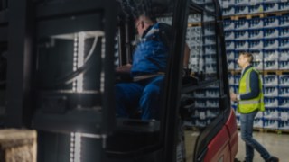 A pedestrian in a high-visibility vest walks through the warehouse behind a forklift truck. A pedestrian in a high-visibility vest walks through the warehouse behind a forklift truck.