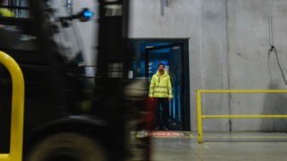 A man in safety clothing steps out of a door into a factory hall. A forklift truck moves past. A man in safety clothing steps out of a door into a factory hall. A forklift truck moves past.