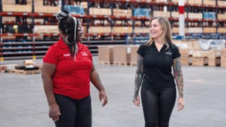 Two female employees dressed in Linde company clothing talk in a modern warehouse environment, symbolising cultural diversity, collegiality and close cooperation in a globally networked, locally rooted team. 