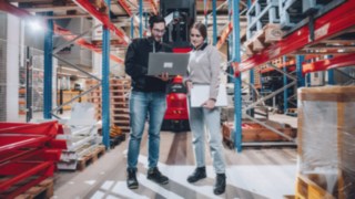 Two employees stand in front of a Linde vehicle in an automated high-bay warehouse and discuss technical matters on a laptop, symbolising work on modern automation solutions in an innovative, technology-driven environment. 