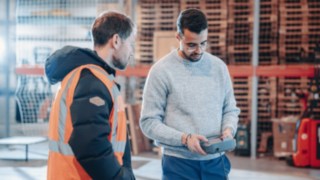 Two employees engaged in technical discussion in a warehouse or industrial environment, one wearing a high-visibility vest and the other operating a control device, symbolising international project assignments, technical responsibility and global career opportunities.