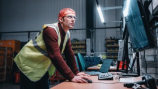 An employee wearing a safety vest works intently at a computer workstation in the hall, symbolising the innovative and technology-driven working environment at KION Automation.