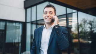 An employee in business attire smiles as he talks on the phone in front of the office building and is actively involved in commissioning at Linde. 