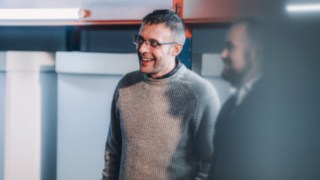 A colleague in a gray knit sweater laughs heartily while talking to a team member as he works in commissioning at Linde. 