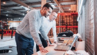 Two colleagues analyse project plans on laptops in a modern warehouse environment, demonstrating creative collaboration at KION Automation.