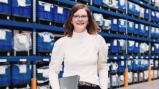 A project manager with a laptop in her hand stands in front of a high-bay warehouse with blue containers – symbolizing responsibility, organizational talent, and strategic work in project management within logistics processes. 