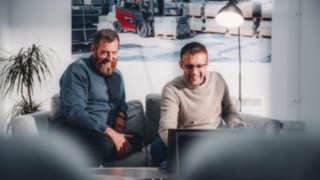 Two employees sit relaxed on a sofa and laugh together at content on a laptop, symbolizing an inspiring work environment that offers space for creativity, innovation, and personal development.