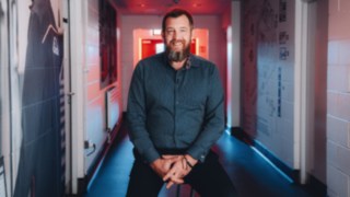 A sales employee sits relaxed in a strikingly lit company corridor and smiles at the camera, representing personality, customer focus and authentic commitment in international sales. 