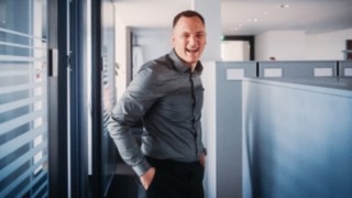 A smiling sales employee in a modern office environment embodies the diversity and dynamism of everyday sales life with constantly new customer contacts, challenges and solutions. 