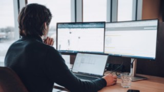 A KION Automation employee works on technical drawings and automation plans at a desk with three screens, symbolizing the use of state-of-the-art technologies in intralogistics. 