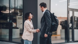 Two businesspeople greet each other with a handshake in front of a modern office building, symbolizing international cooperation, cultural openness, and global career opportunities within a networked corporate environment.