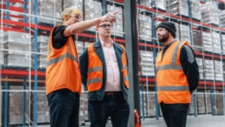 Three employees in high-visibility vests are having a conversation in a modern high-bay warehouse, embodying team-oriented cooperation, professional exchange, and a collaborative corporate culture in a dynamic working environment.