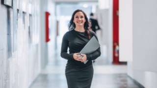 A confident employee with a laptop in her hand walks smiling through a modern office corridor, representing dedicated work on projects that make global supply chains more efficient and sustainable.