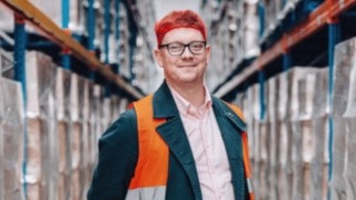 An employee from the Solution Design department at KION Automation, wearing a high-visibility vest and red hairnet cap, stands professionally and friendly in a high-bay warehouse. 