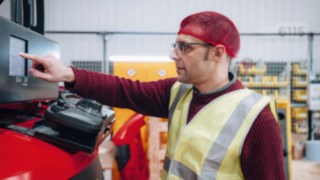 An employee from KION Automation's commissioning department, wearing a reflective vest and a red cap, configures a Linde vehicle in a high-bay warehouse. 
