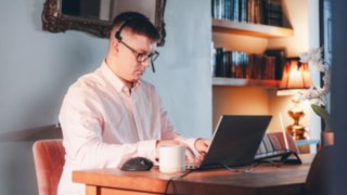 A man is sitting at a desk, staring intently at a screen. He is wearing a headset with a microphone. He is wearing a dark blue jumper with a light geometric pattern and a light blue shirt underneath.